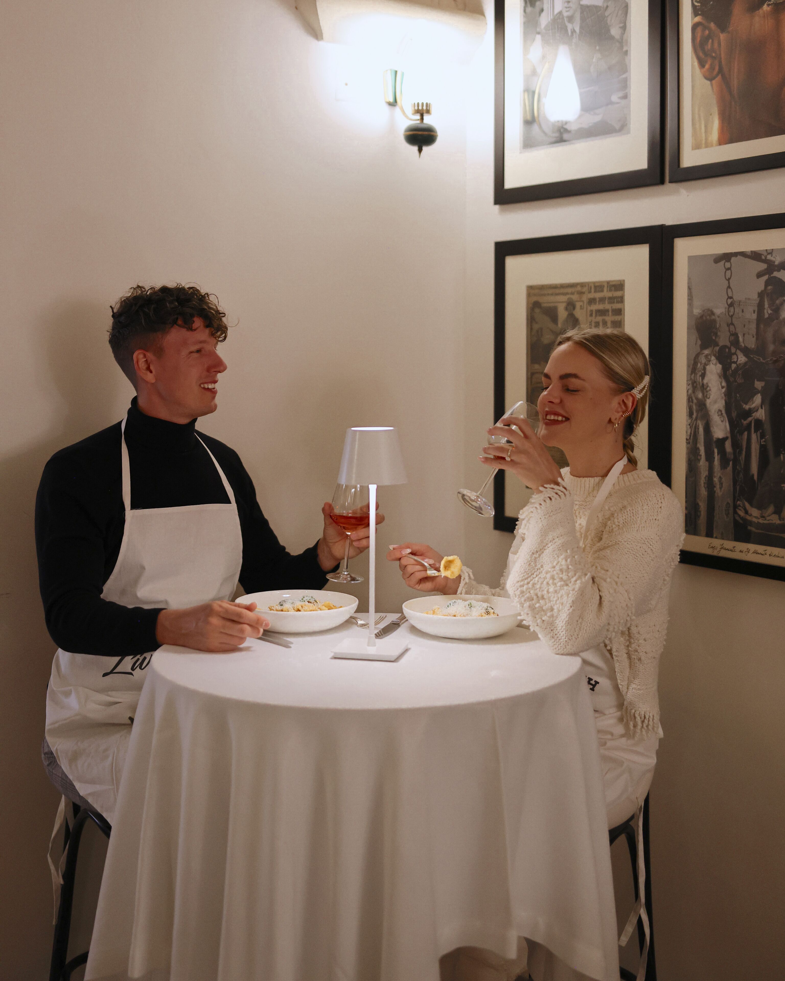 Two people dining at a round table with white tablecloths. There is a lamp on the table, and the walls are adorned with framed pictures.