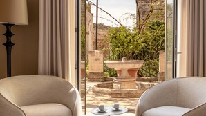 Una habitación con dos sillones color beige frente a una mesita con dos tazas, un ventanal grande y vistas a un patio con una fuente y follaje.
