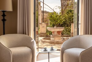 Una habitación con dos sillones color beige frente a una mesita con dos tazas, un ventanal grande y vistas a un patio con una fuente y follaje.