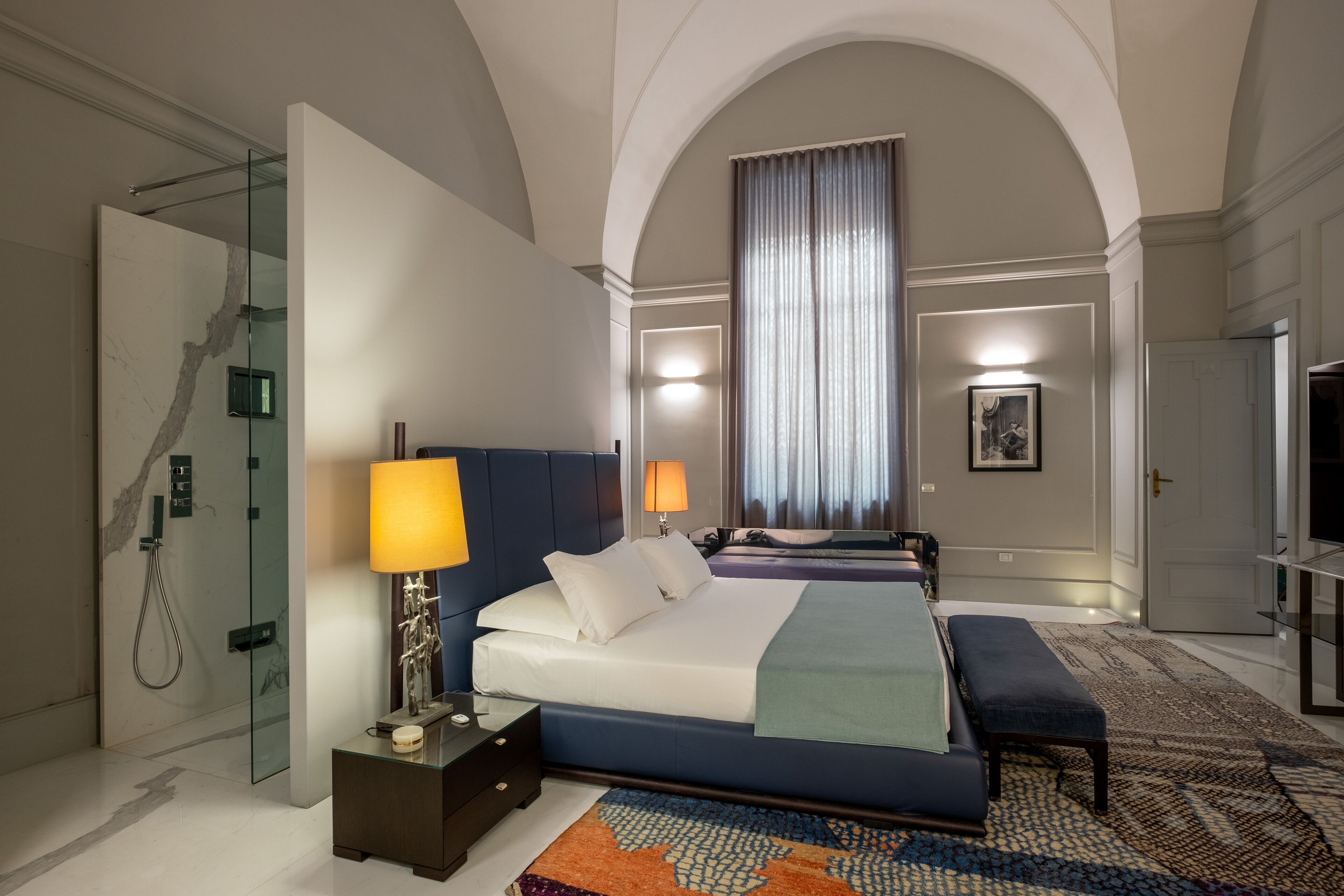 Een moderne hotelkamer met een groot bed, een bankje, een douchecabine van glas en uitzicht op de stad door een groot raam.