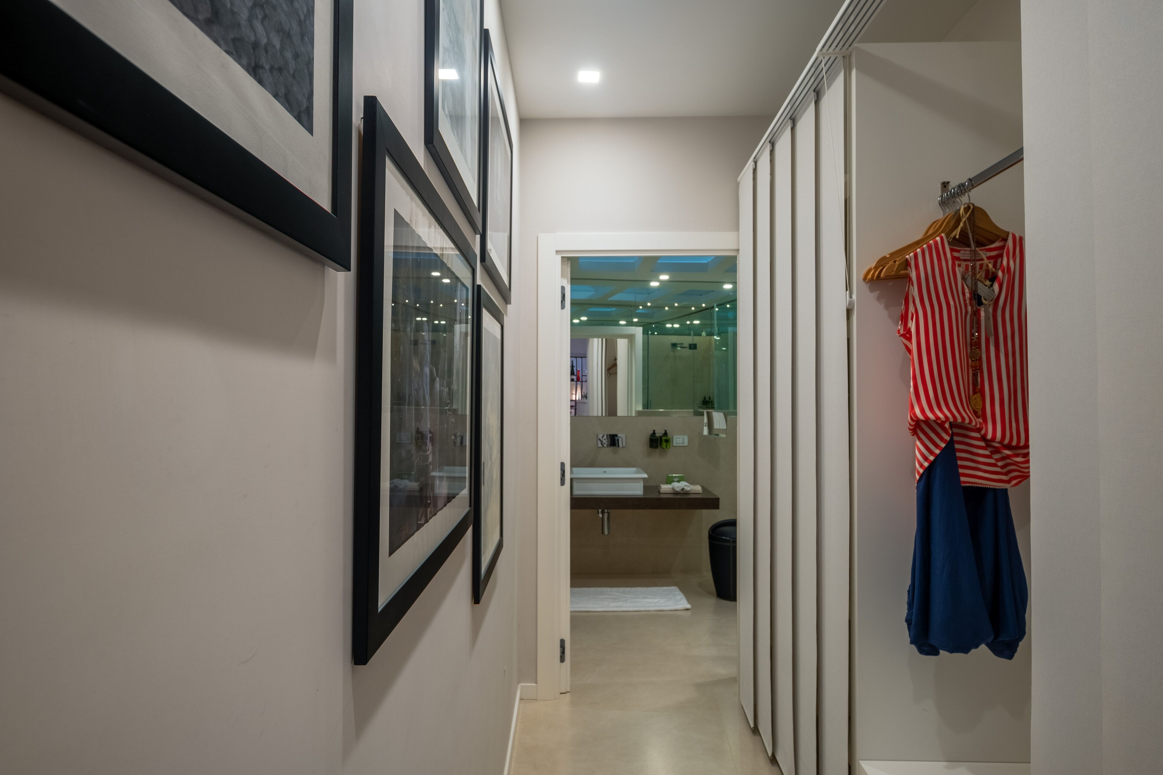 Un couloir étroit, avec des tableaux encadrés sur le mur de gauche, une salle de bain avec un lavabo et un miroir sur le mur de droite, et un portant à vêtements sur lequel sont suspendus un t-shirt rayé et une jupe bleue.