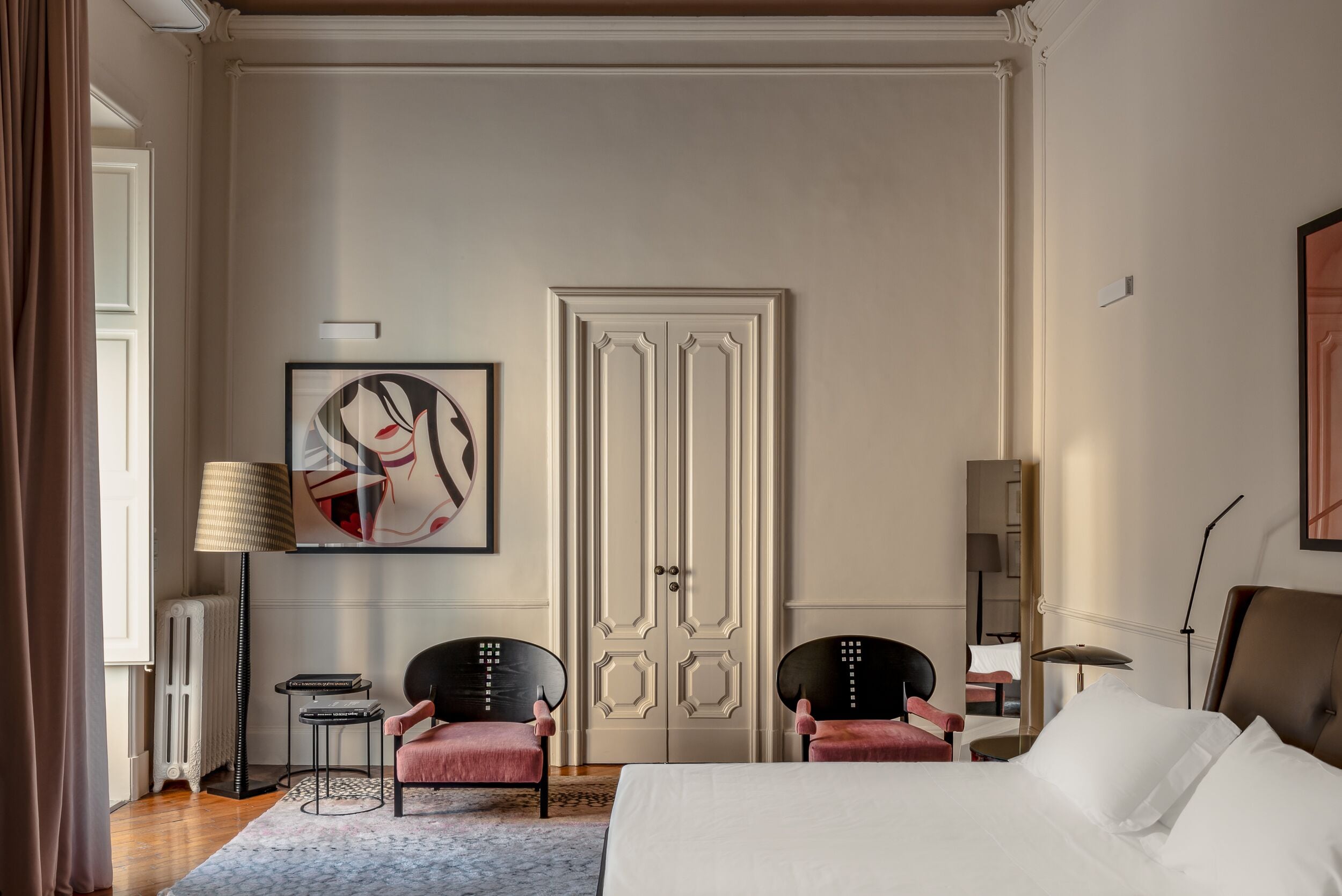 Et hotelværelse med en stor seng, to stole, en radiator, et indrammet kunstværk og en dør med dekorative panelger.'
