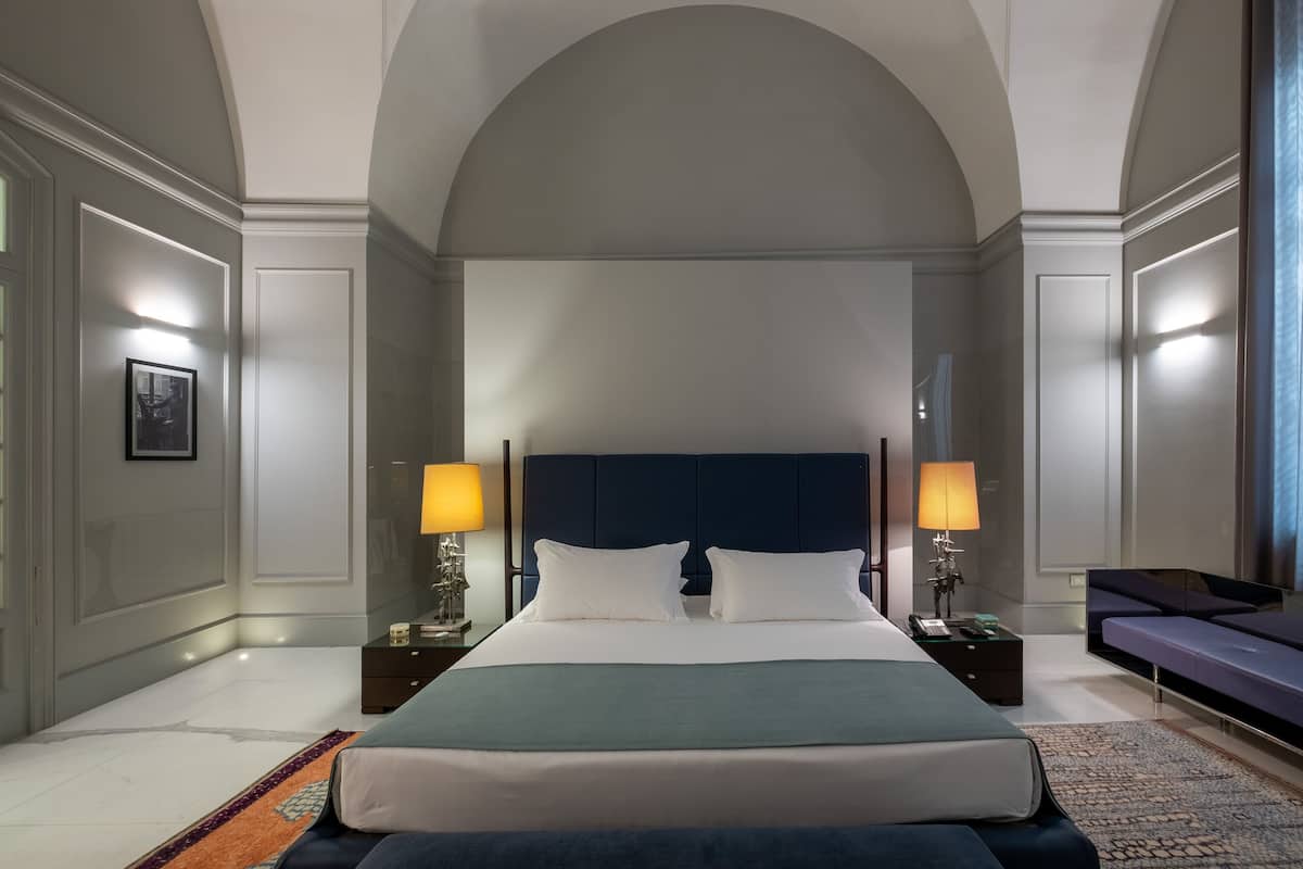 Een moderne hotelkamer met een groot bed, twee nachtkastjes met lampen, een sofa en een hoge, gebogen plafond.