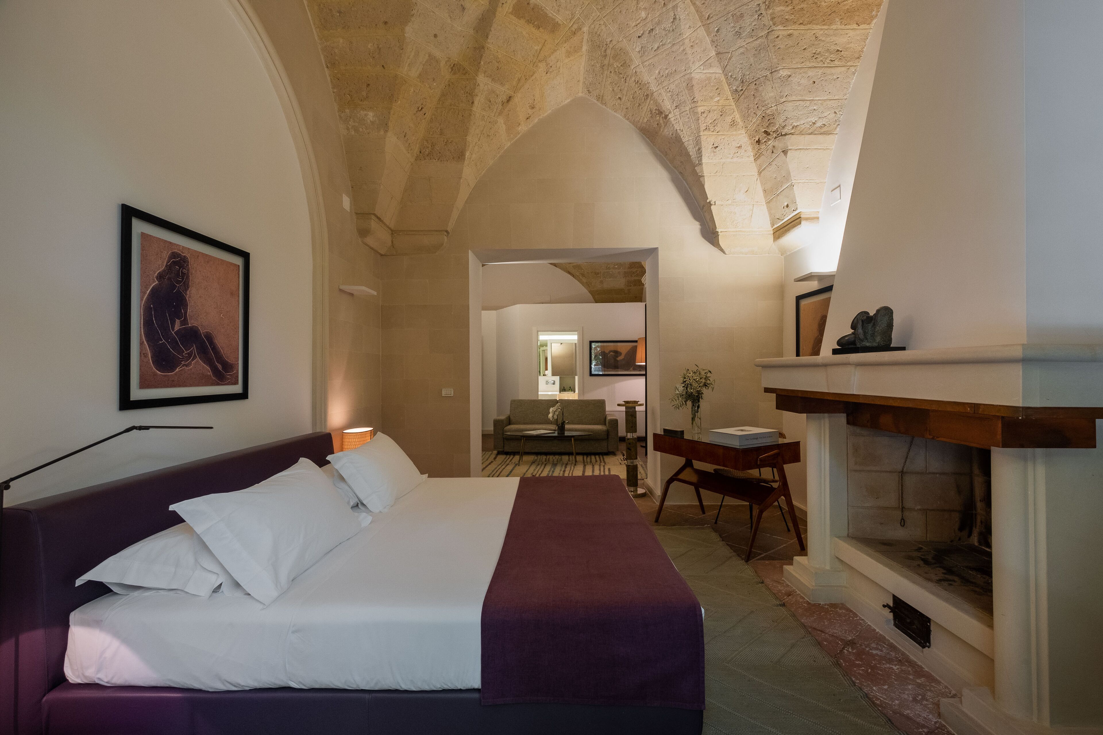 A modern hotel room with a bed, a purple sofa, a fireplace, and a framed artwork on the wall.