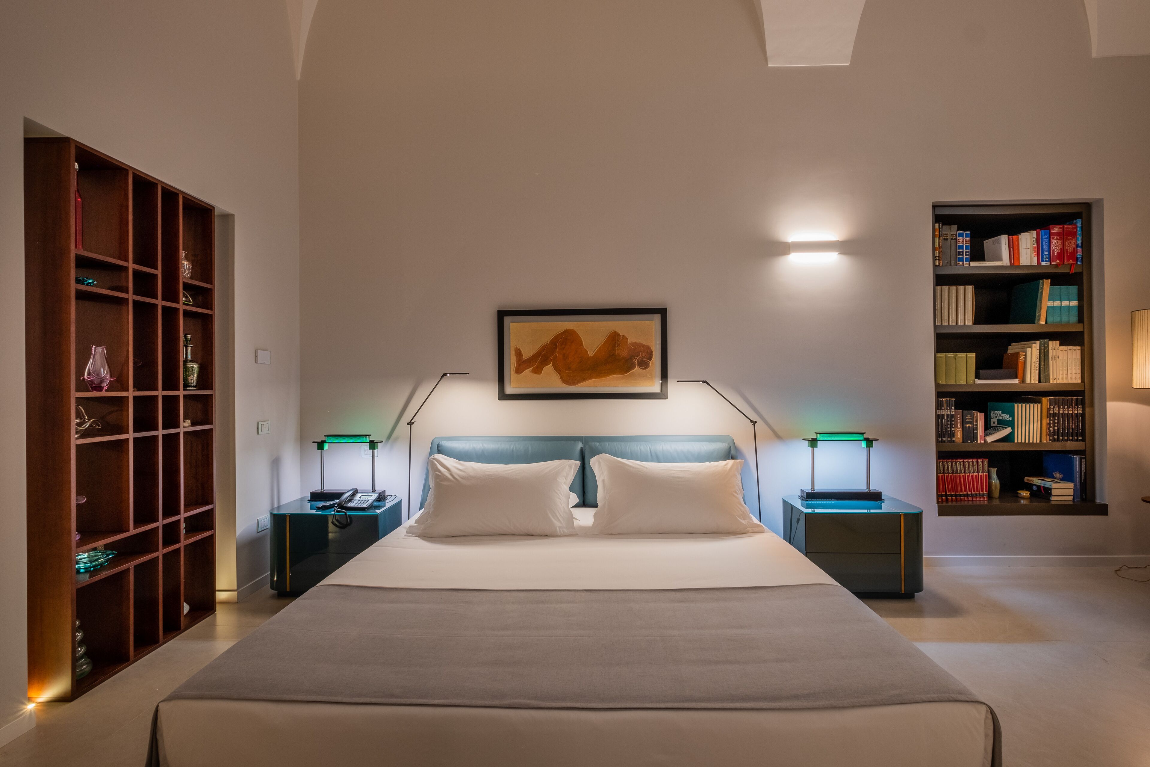 A modern bedroom with a large bed, a wooden bookshelf, a desk with a lamp, and a wall-mounted bookshelf.