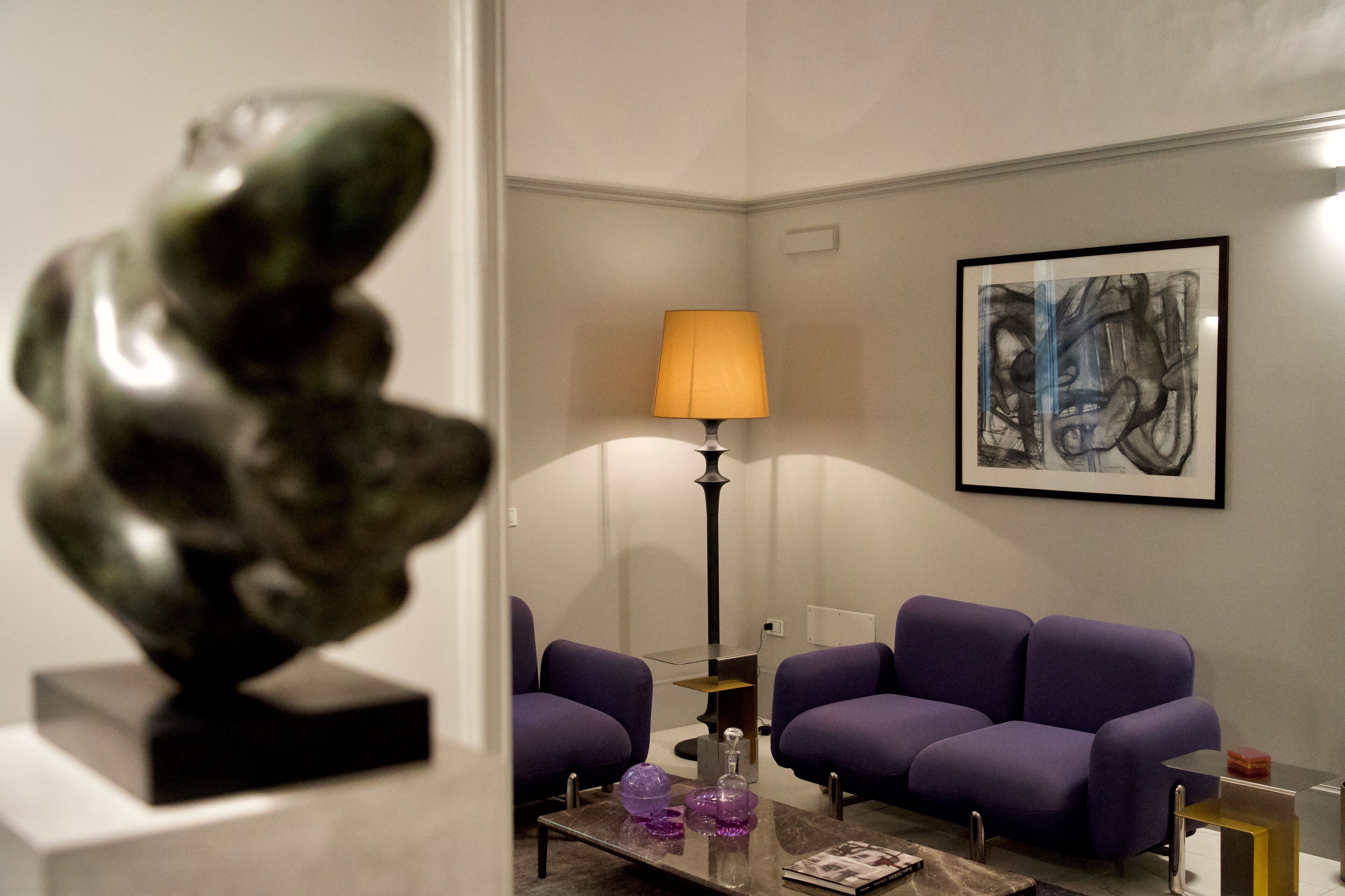 Un salon moderne avec des canapés violets, une table basse en verre, une lampe et une sculpture.