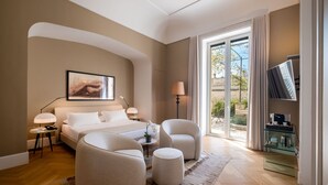 Una habitación de hotel moderna con una cama grande, dos sillones, una mesita y un televisor sobre un mueble de vidrio.