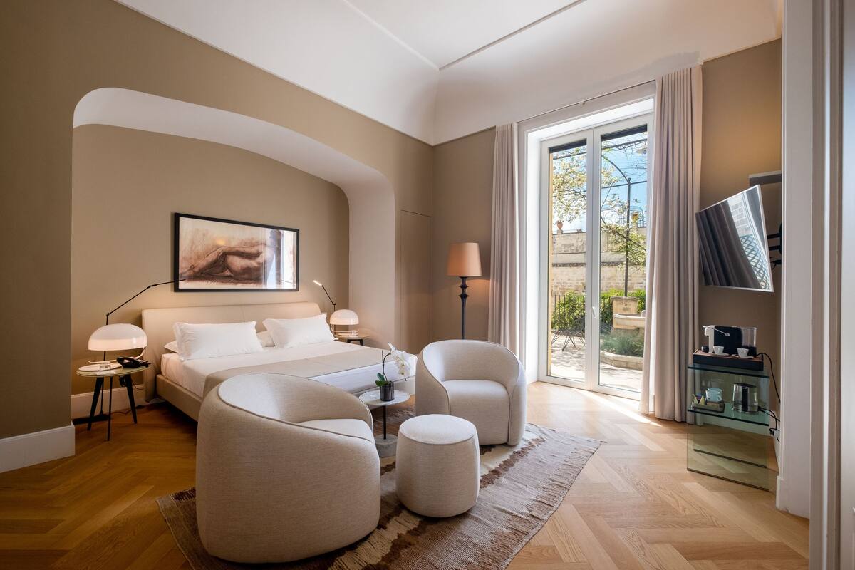 Een moderne hotelkamer met een groot bed, twee fauteuils, een klein tafeltje en een televisie op een glazen kast.