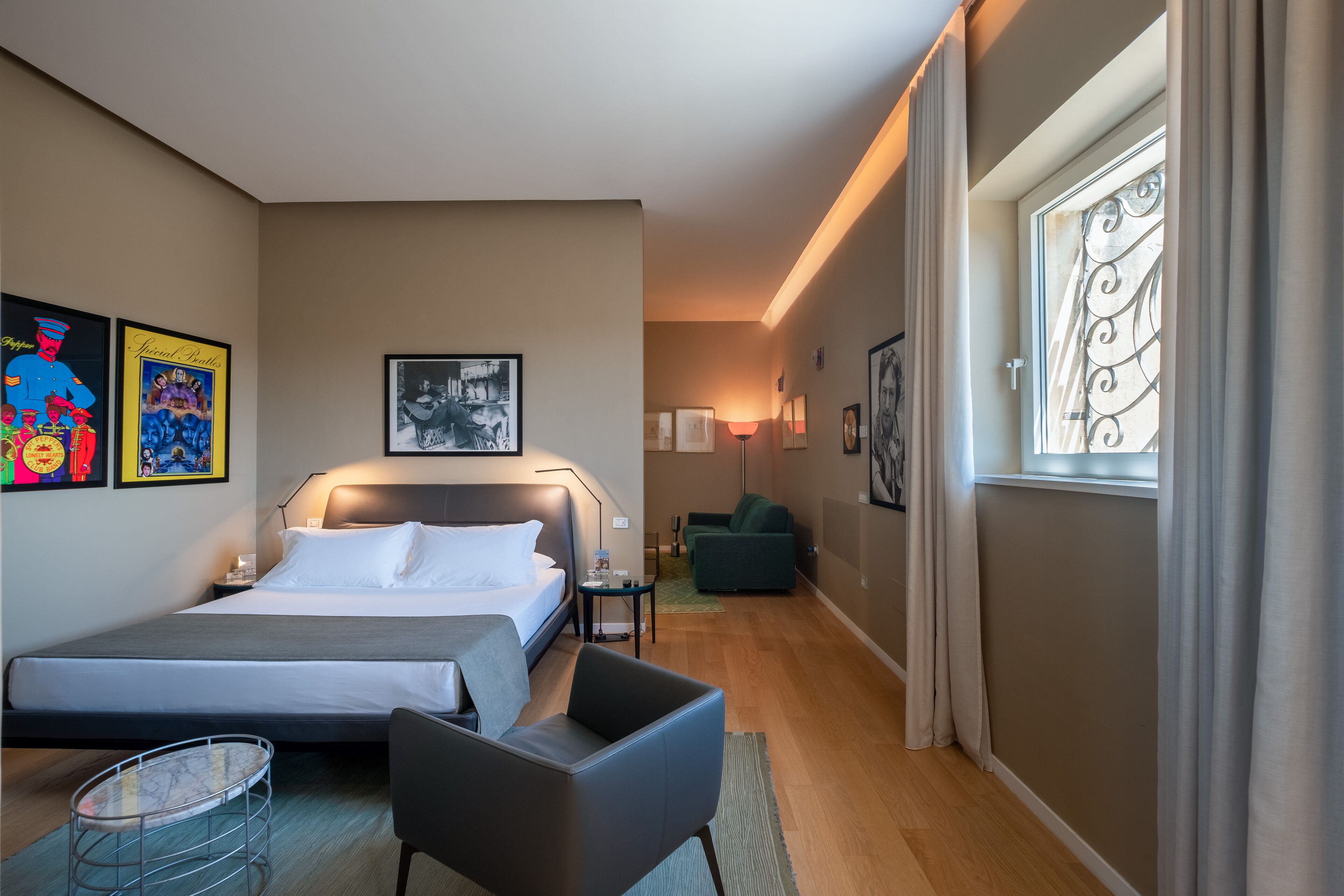 A modern hotel room with a large bed, a desk, a chair, and a small table. There is a window with curtains, a lamp, and artwork on the walls.