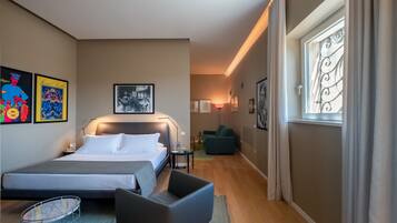A modern hotel room with a large bed, a desk, a chair, and a small table. There is a window with curtains, a lamp, and artwork on the walls.