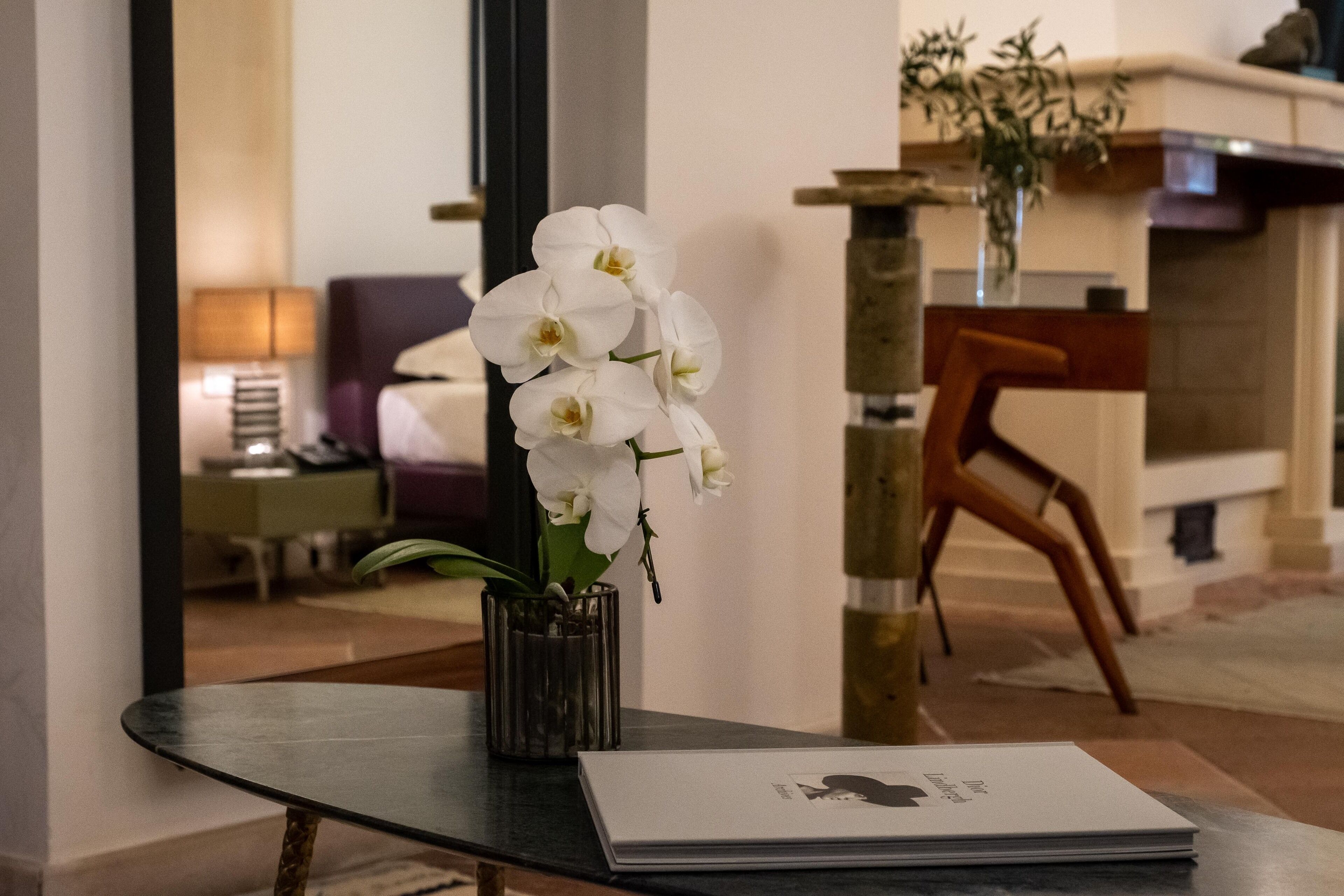 A reception desk with a vase of white orchids, a magazine, and a mirror reflecting a room with a lamp and a chair.