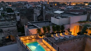 Un área de piscina en la azotea con vistas a la ciudad al atardecer, que incluye una piscina, sillas de descanso y plantas en macetas.