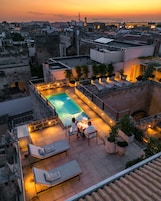 Un área de piscina en la azotea con vistas a la ciudad al atardecer, que incluye una piscina, sillas de descanso y plantas en macetas.