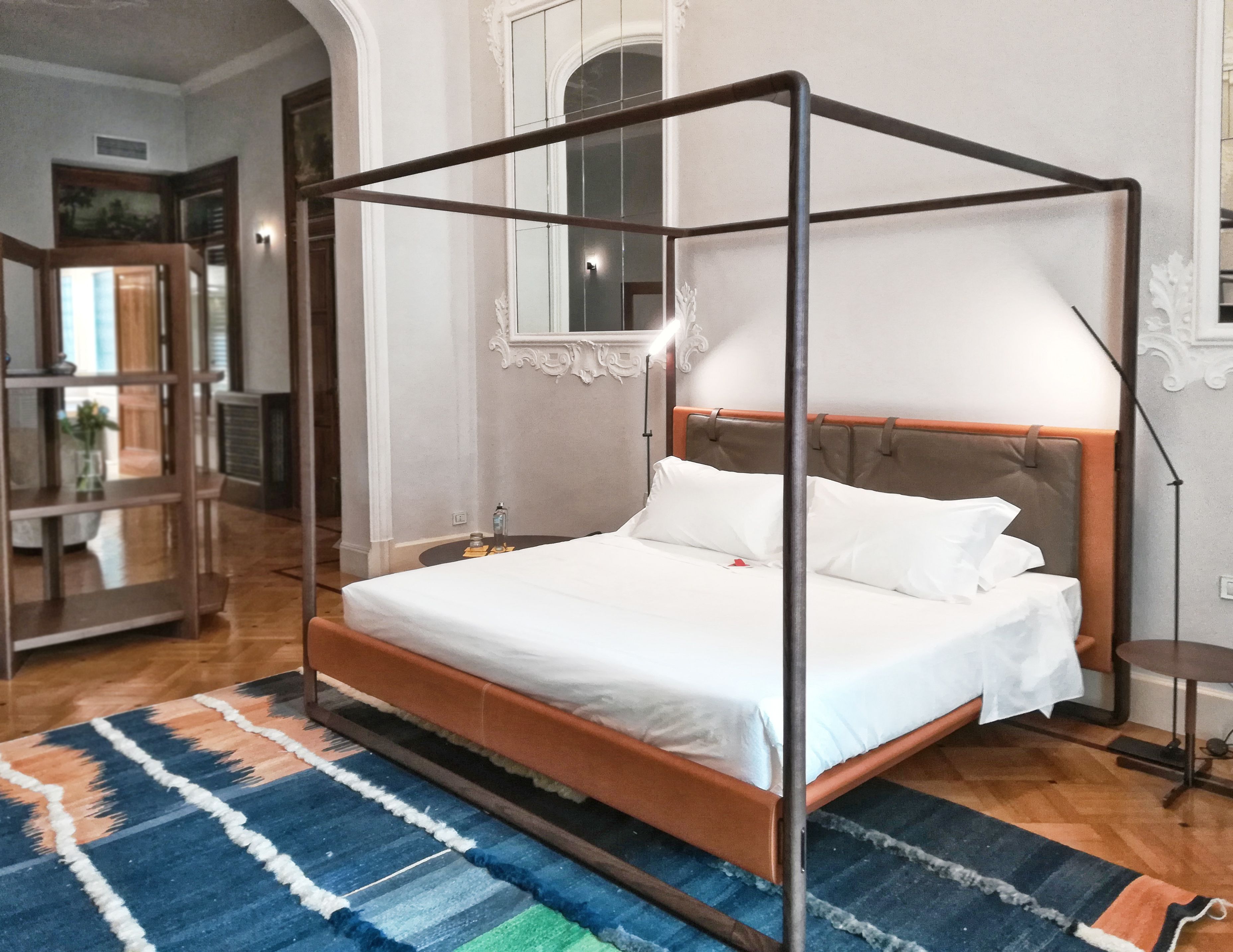 A four-poster bed with a wooden frame and a headboard, a white bedspread, and a blue and orange patterned rug.