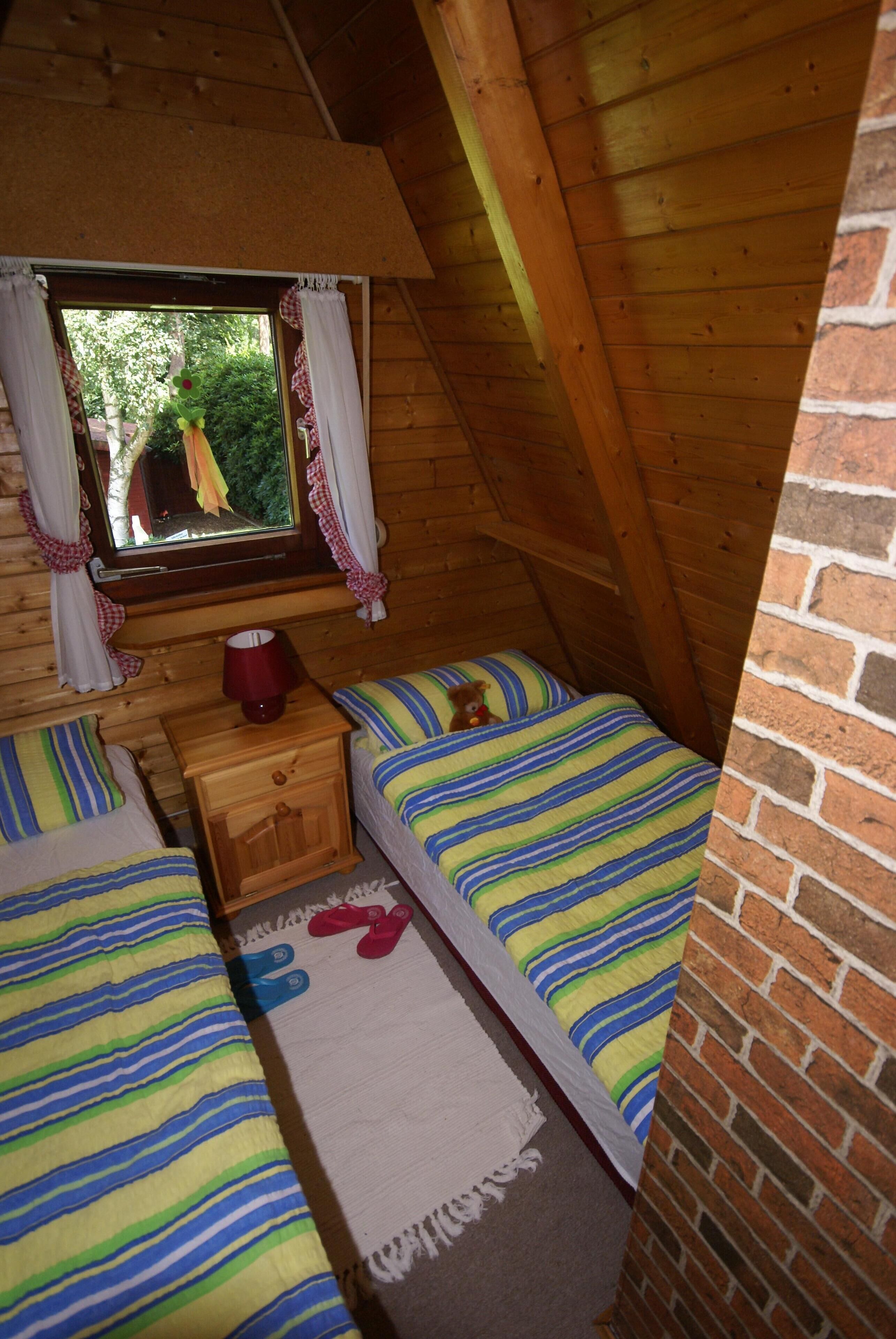 A cozy attic bedroom with two beds, a window with curtains, a wooden nightstand, and a brick wall.