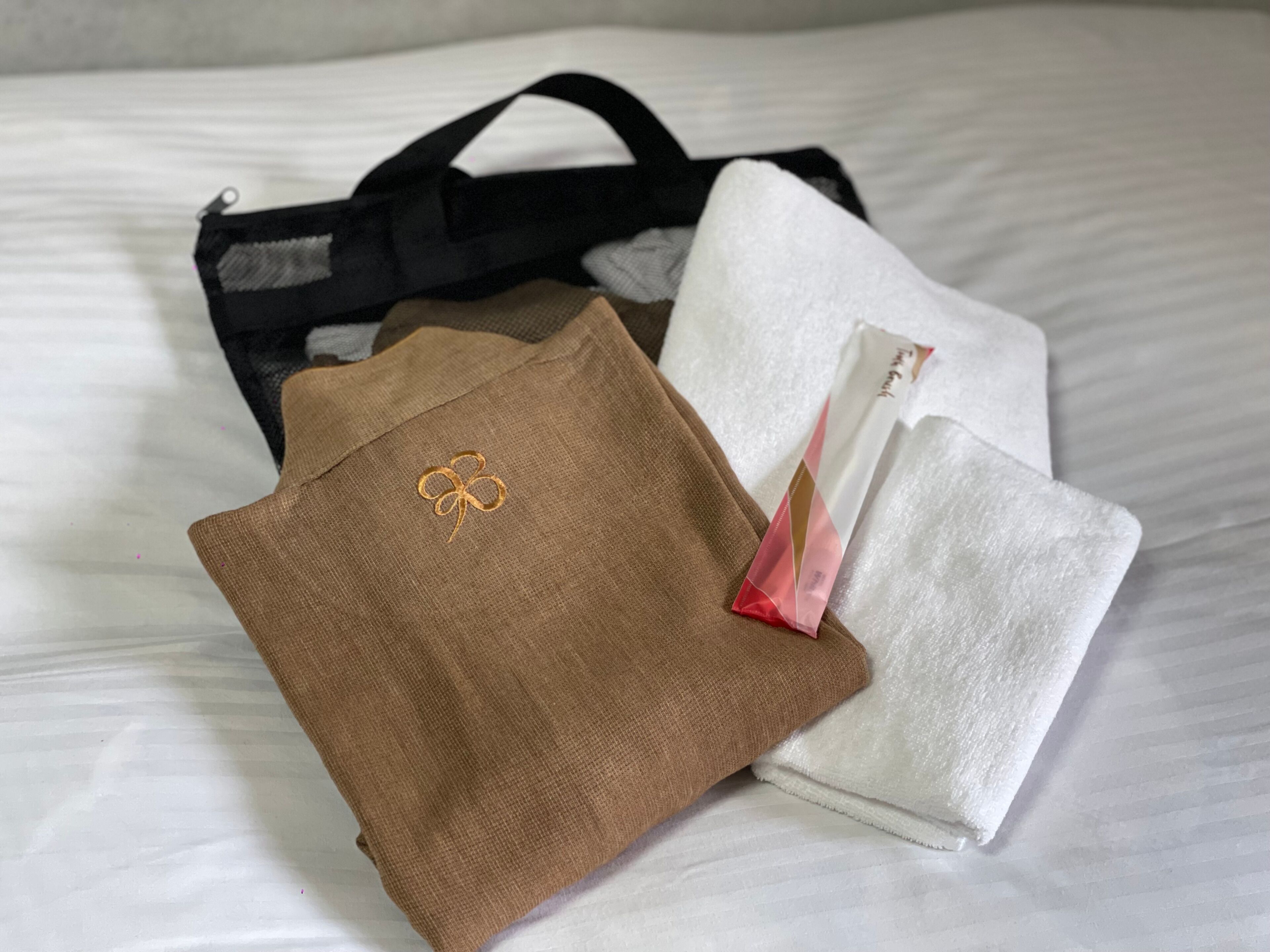 Una cama con sábana blanca, almohada marrón con diseño de trébol y una bolsa negra.
