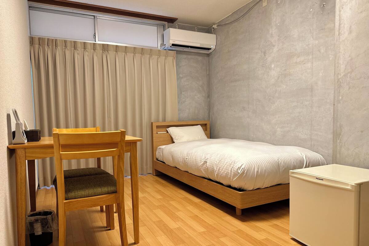 Ein Schlafzimmer mit einem hölzernen Bett, einem kleinen, weißen Kühlschrank, einem Holztisch mit Stuhl und einem Fenster mit Vorhängen.