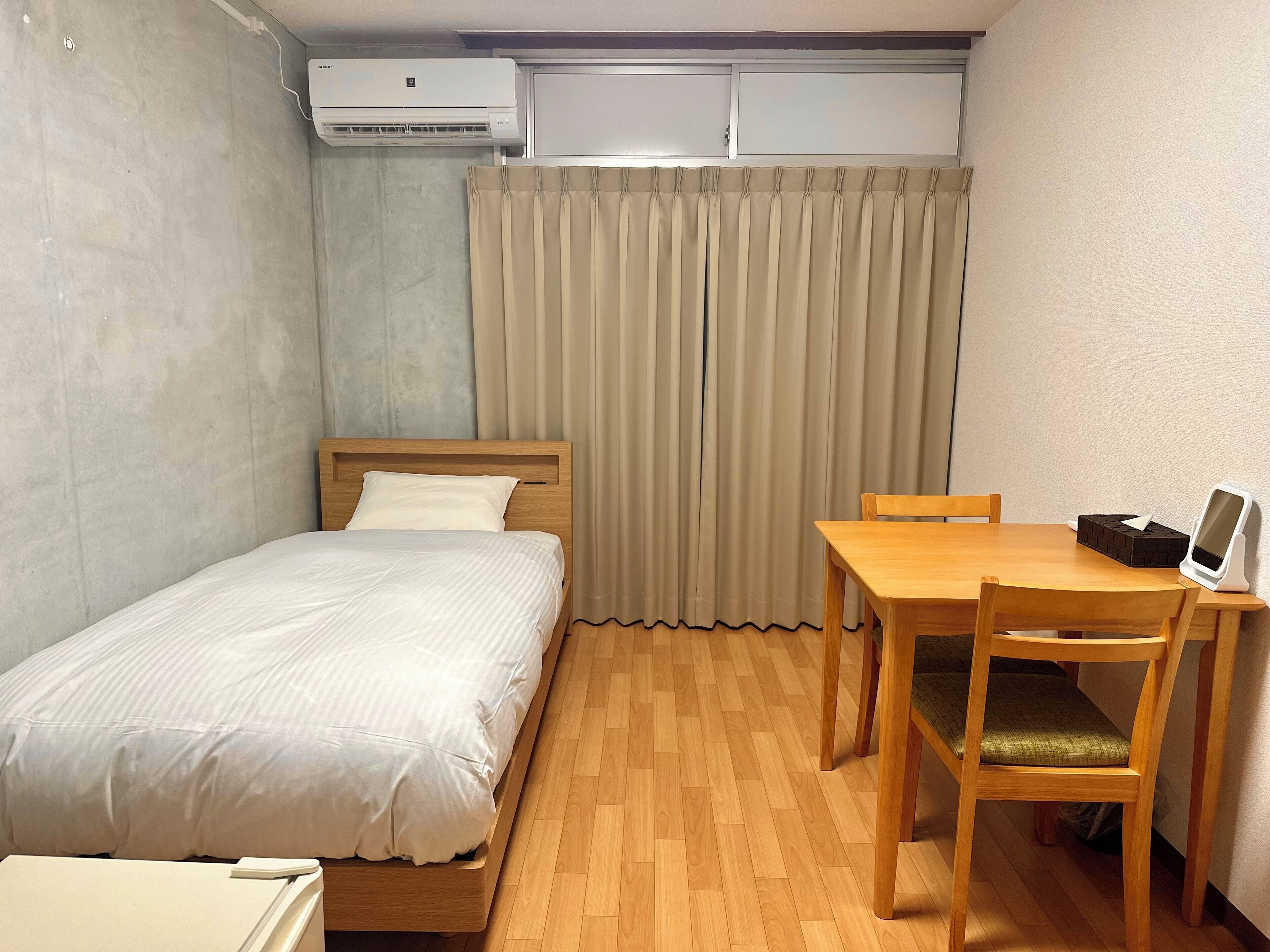 A small, neatly arranged room with a bed, a table, and a chair. There is an air conditioning unit above the bed and a curtain covering the window.