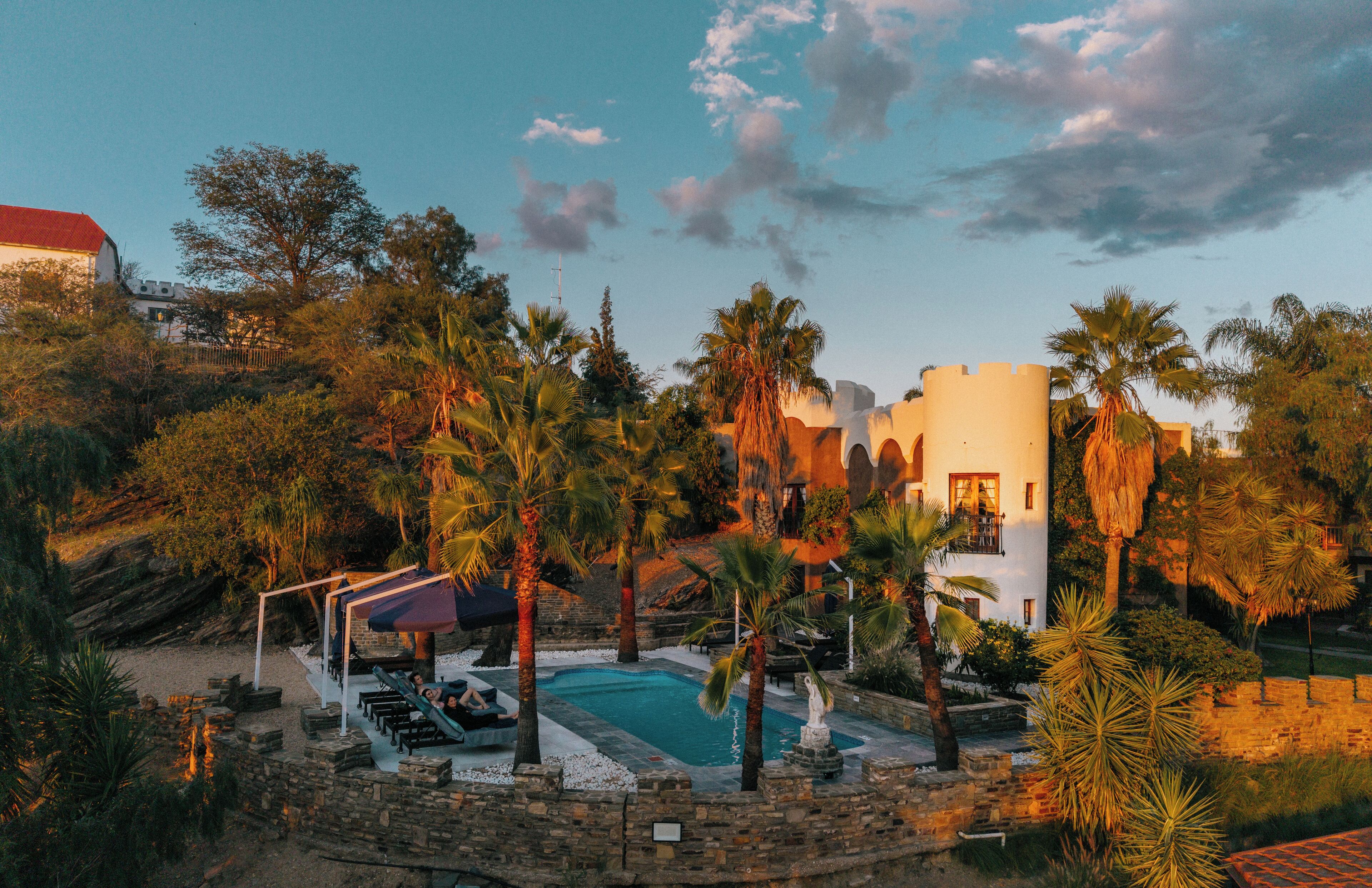 Zona de piscina con sillones de descanso, palmeras y un edificio con techo rojo.