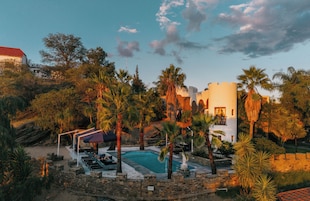 Zona de piscina con sillones de descanso, palmeras y un edificio con techo rojo.