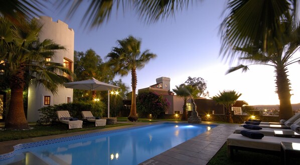 A swimming pool with lounge chairs and umbrellas, surrounded by palm trees and a building with a tower.
