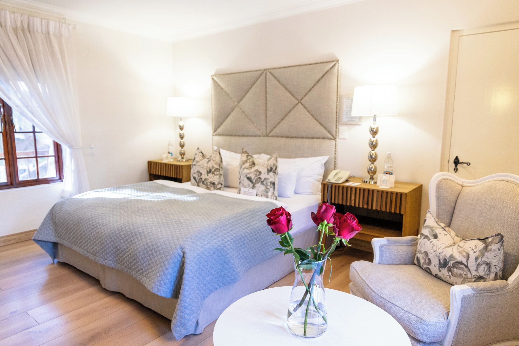 Ett hotellrum med en stor säng, ett nattduksbord med en lampa, en stol och en vas med blommor.