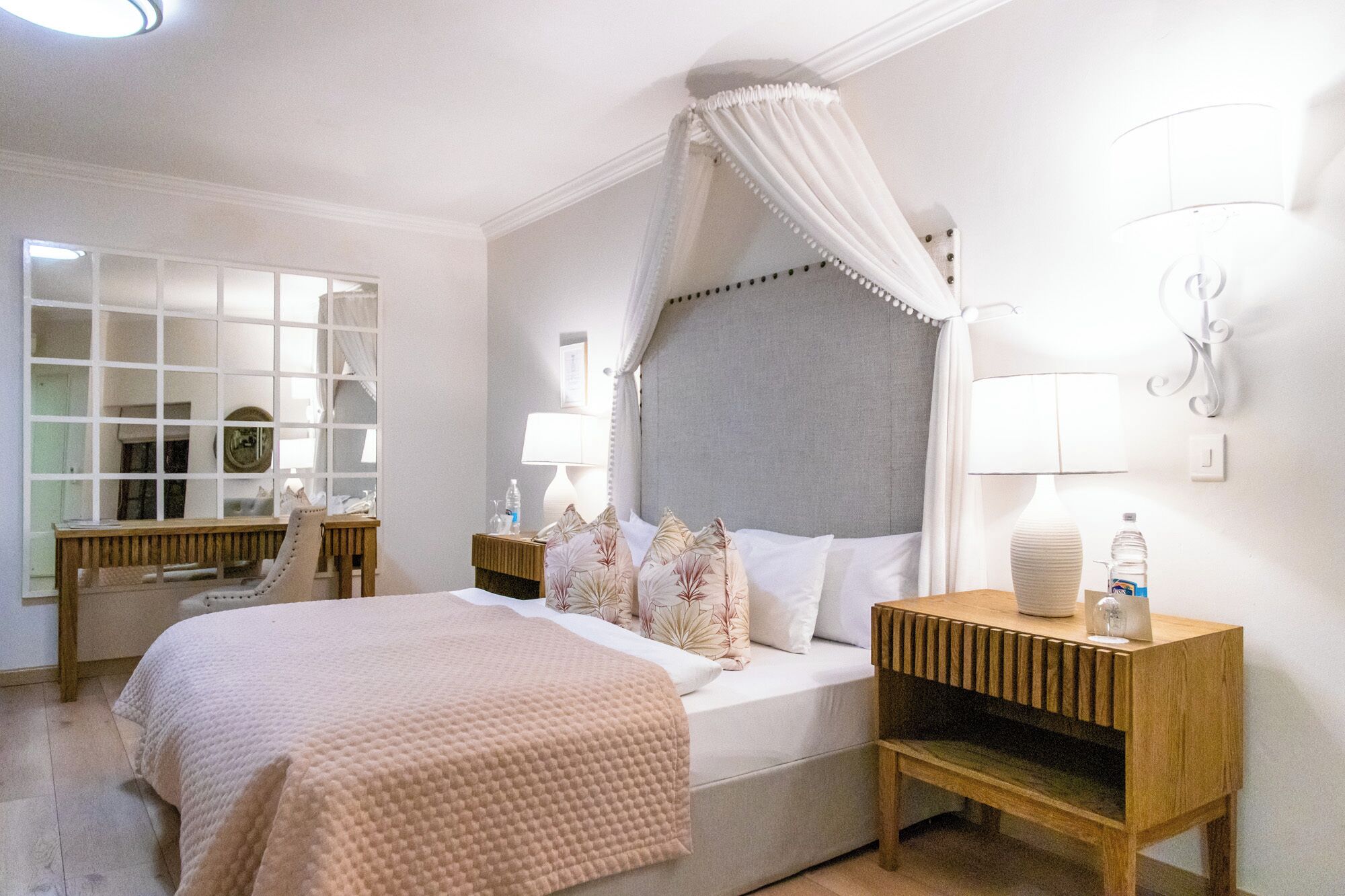A hotel room with a large bed, a bedside table with a lamp, a mirror, and a view of the outside.