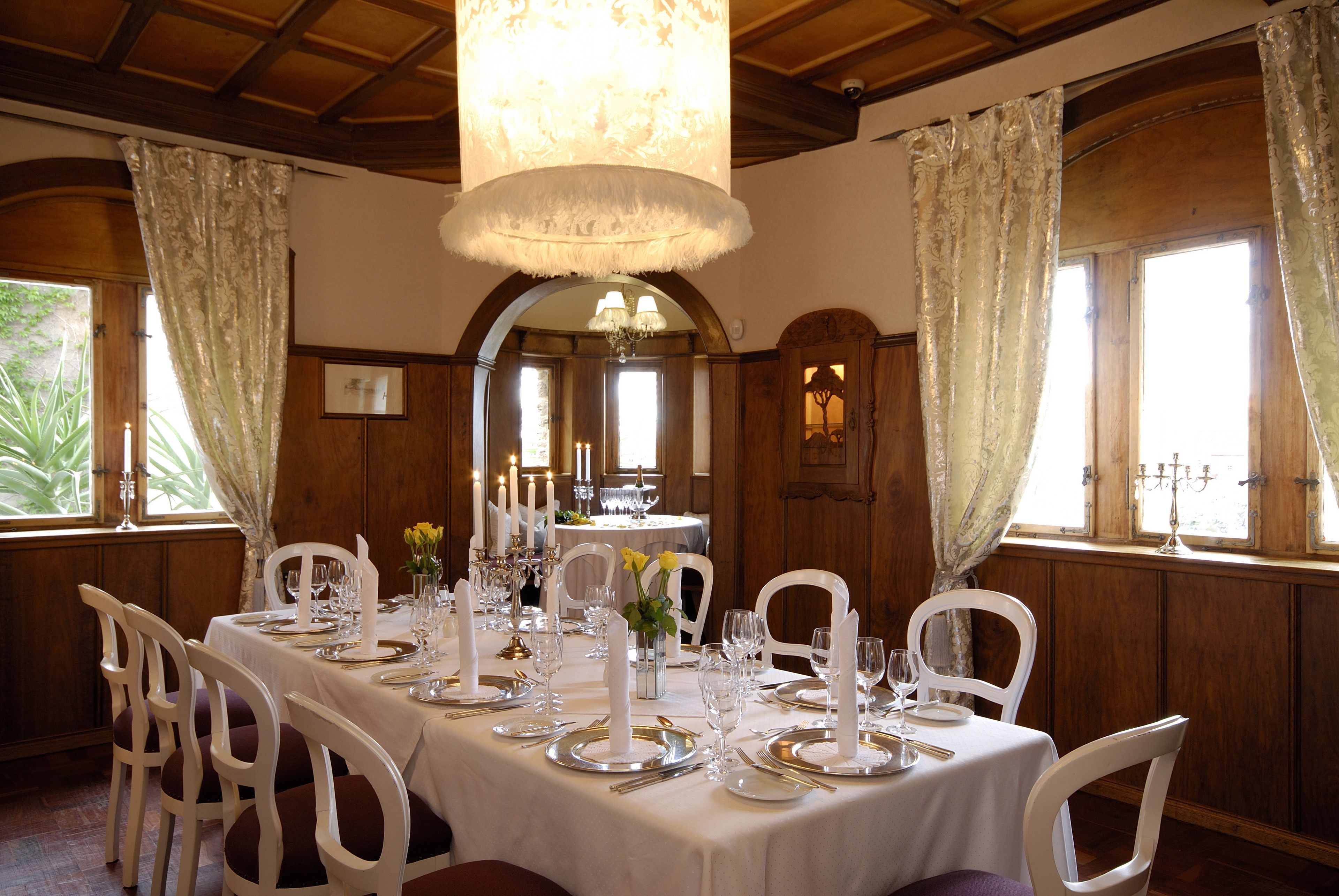 A dining room with a long table set for a meal, surrounded by white chairs, with a chandelier above and wooden paneled walls.