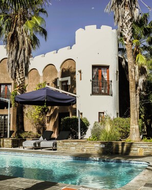 A pool area with lounge chairs and umbrellas, a white building with arches, and palm trees.