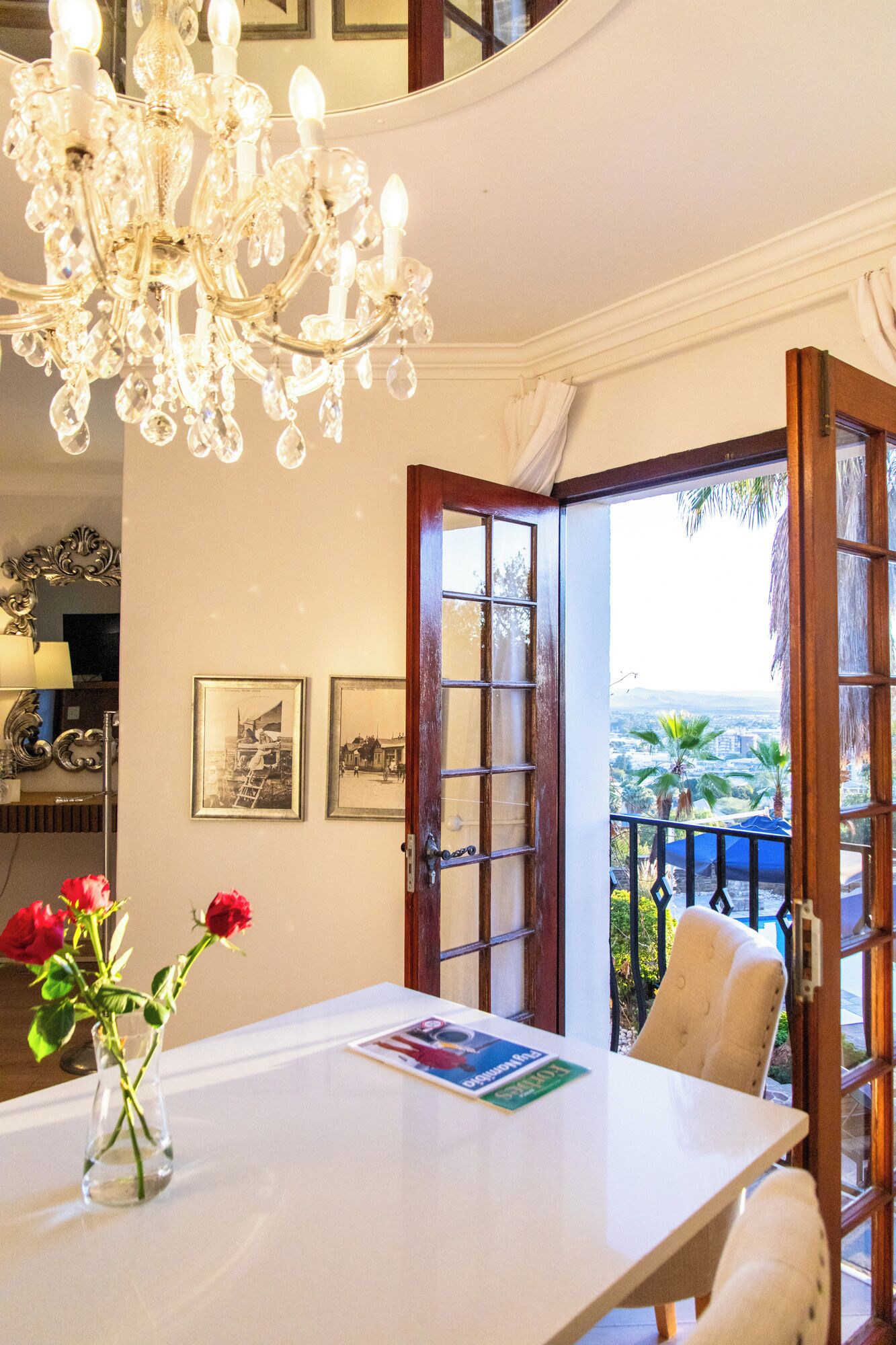 A dining area with a chandelier, a table with a vase of red roses, and a view of the outside through open doors.