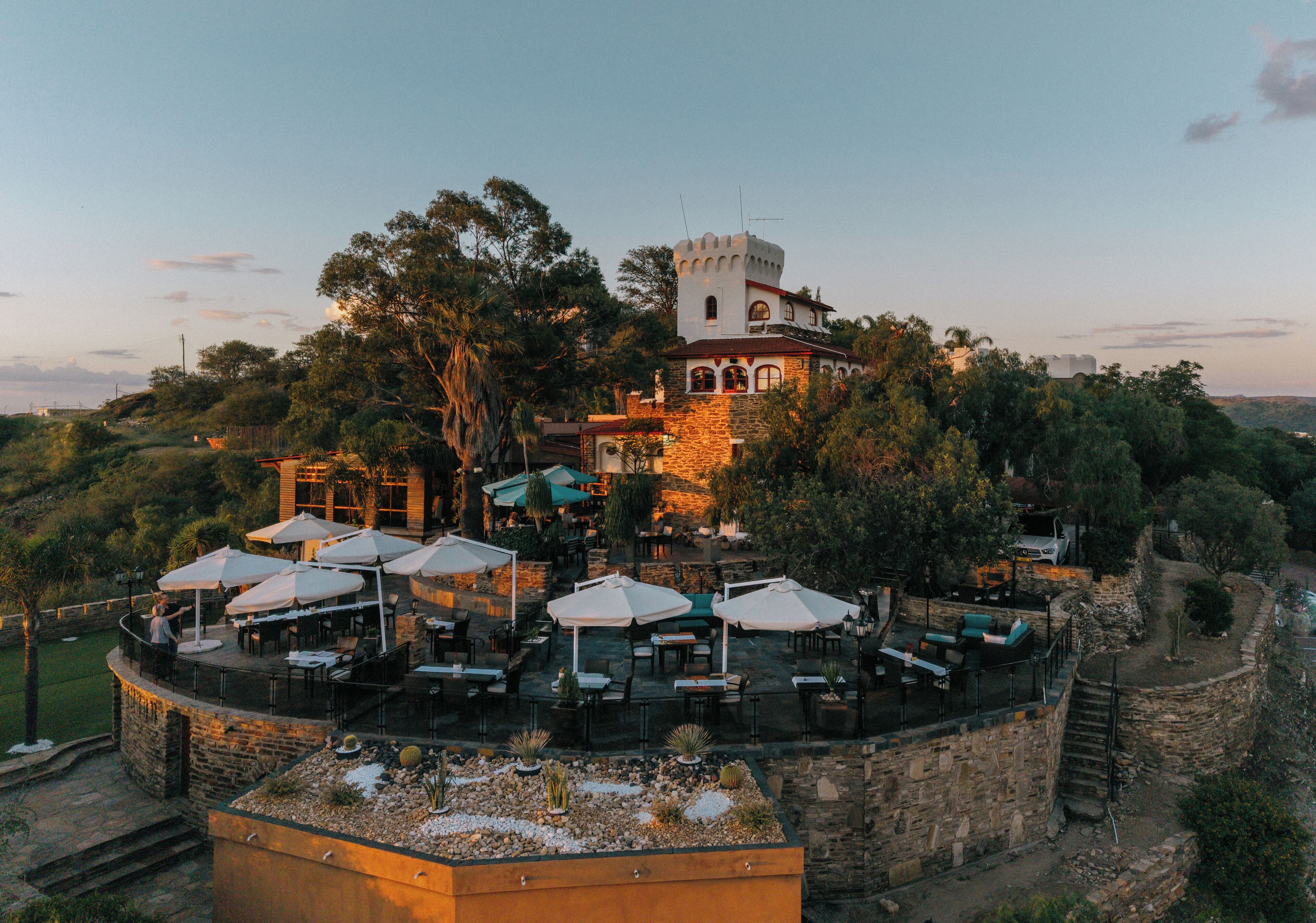 A castle-like building with a tower, surrounded by lush greenery and outdoor seating with umbrellas.
