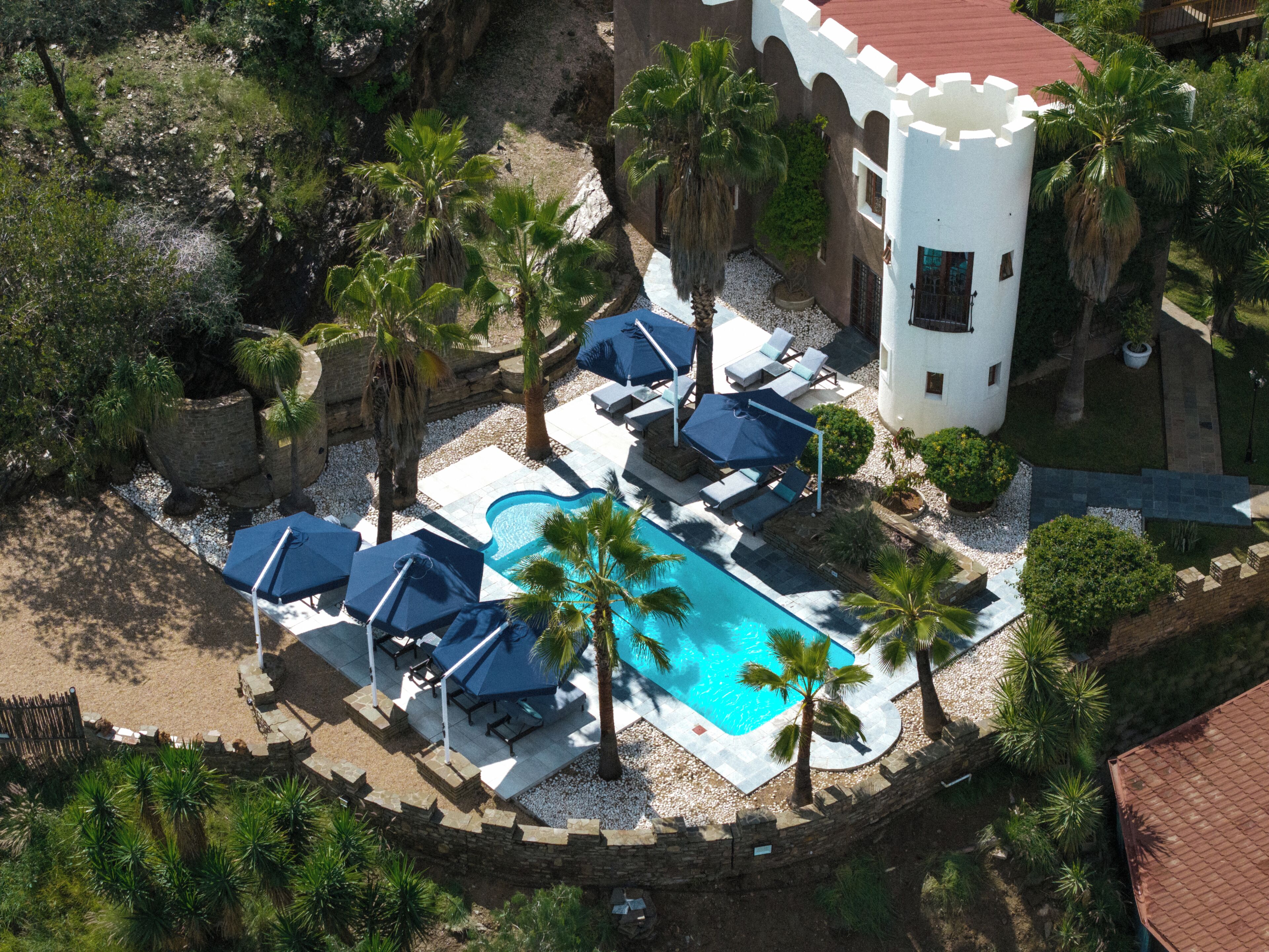 Vista aérea de un resort con piscina, sillas de descanso, palmeras y un edificio parecido a un castillo.