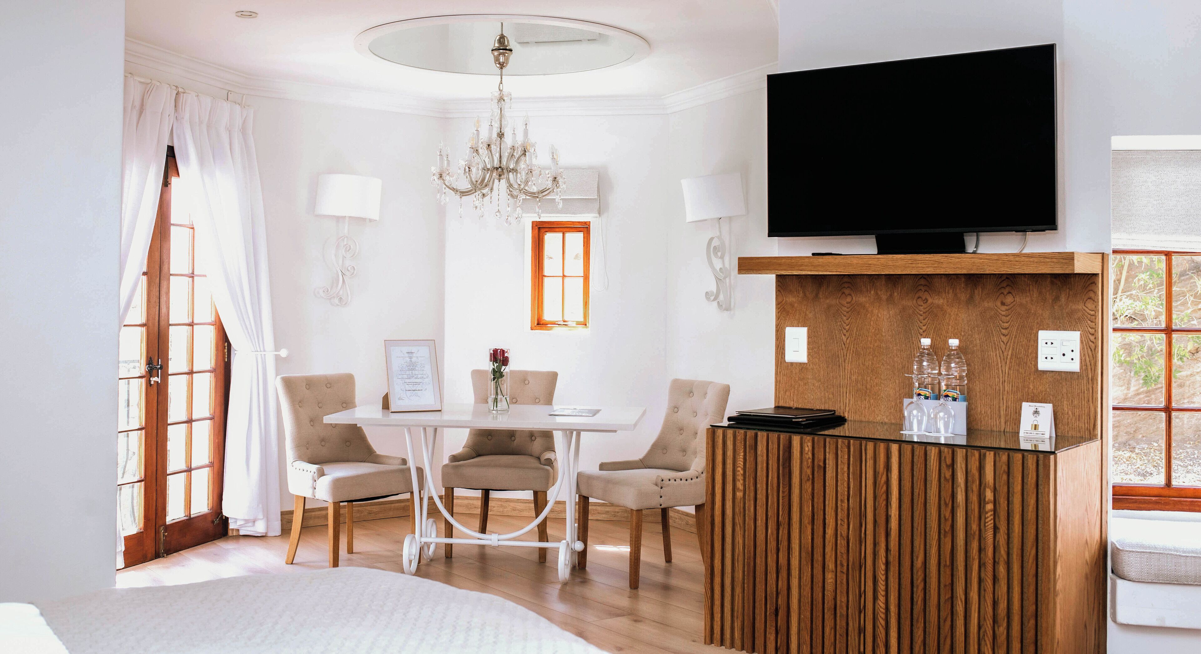 A hotel room with a bed, a dining table with chairs, a TV mounted on the wall, and a wooden reception desk.