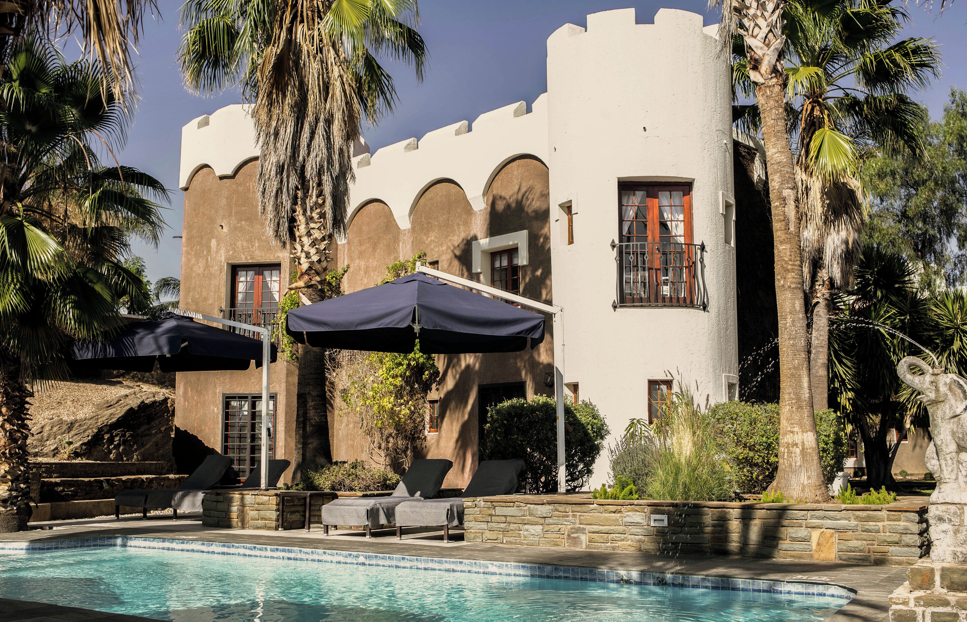 A pool area with lounge chairs and umbrellas, a stone wall, and a building with arched windows and a palm tree.