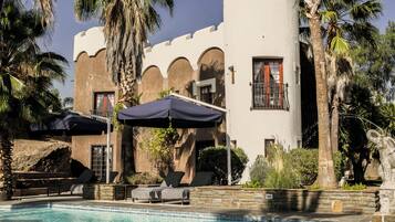 A pool area with lounge chairs and umbrellas, a stone wall, and a building with arched windows and a palm tree.