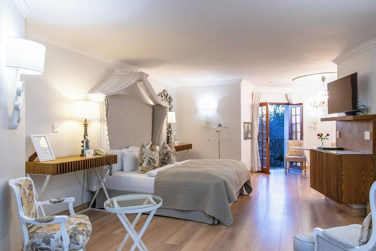 A hotel room with a bed, bedside tables, a chair, a small table, a TV, and a view of the outdoors.