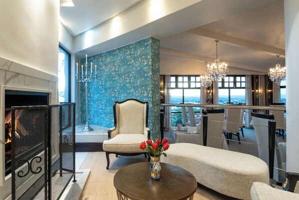 Un salon moderne doté d’une cheminée, d’une grande fenêtre offrant une vue sur la ville, d’un lustre et d’un coin salon agrémenté d’un vase de roses rouges.