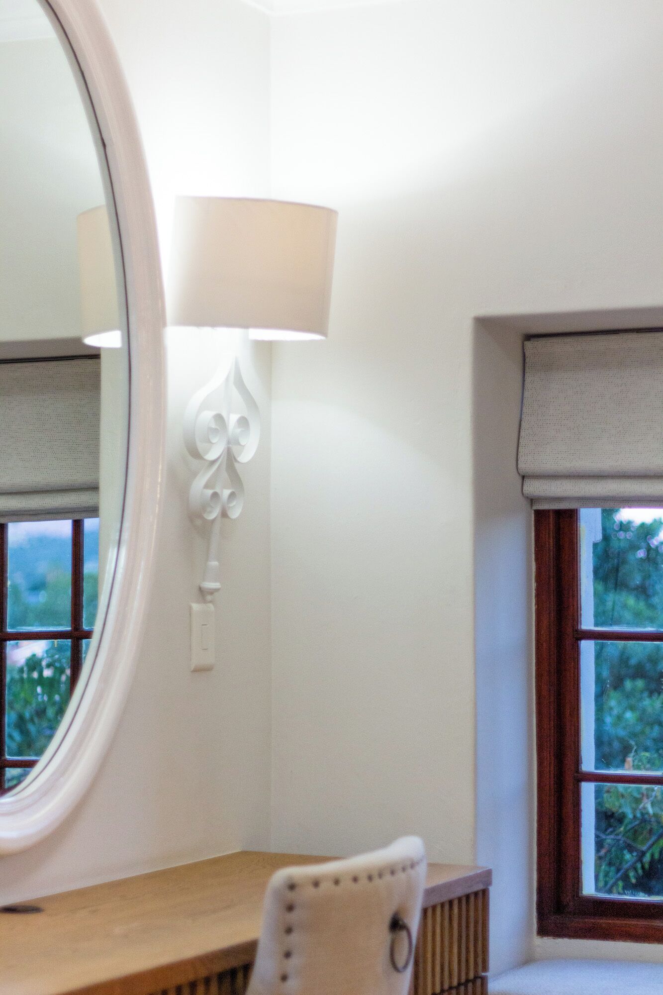 A room with a round mirror, a lamp, a chair, and a window with a view of greenery.
