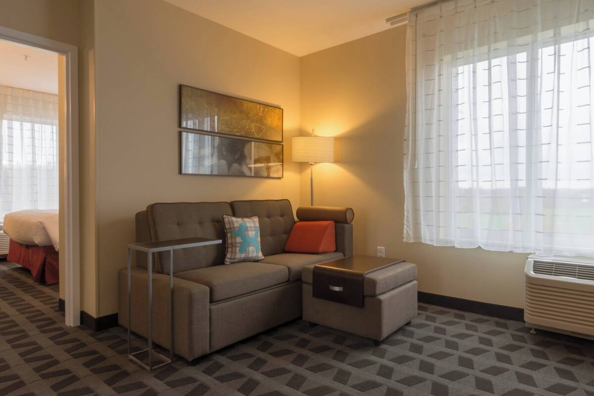 A hotel room with a sofa, a lamp, a window with sheer curtains, and a radiator.