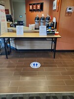 Zona de recepción de un hotel con un mostrador, un letrero que indica distanciamiento social, una pantalla digital y un cartel en la pared con los iconos de Stay, Work y Play.