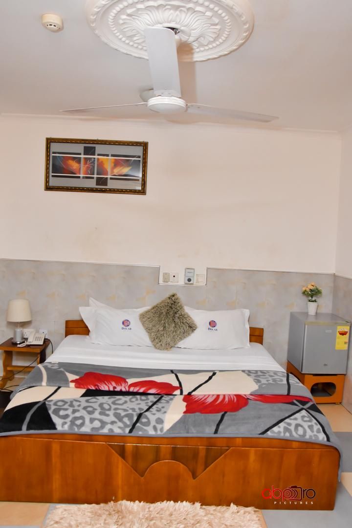Habitación de hotel con cama, ventilador de techo, mesita de noche, lámpara, un cuadro en la pared y una pequeña planta en maceta.