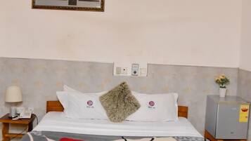 Habitación de hotel con cama, ventilador de techo, mesita de noche, lámpara, un cuadro en la pared y una pequeña planta en maceta.