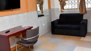 A room with a desk, a black sofa, a chair, a television, and a window with curtains. - Dikab Hotel (Accra)