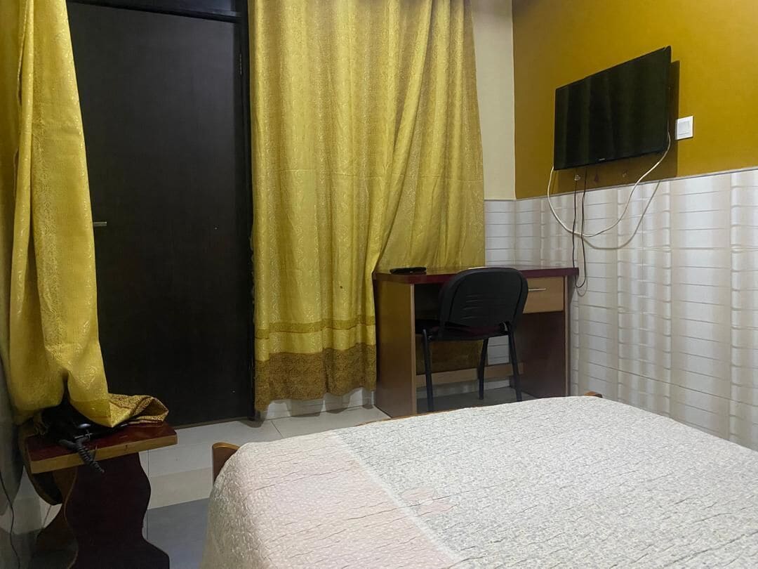 A room with a bed, a desk, a chair, a television mounted on the wall, and a door with curtains.