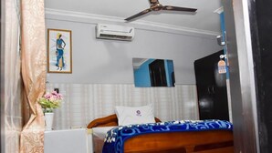 A bedroom with a bed, a ceiling fan, an air conditioner, a framed picture, a vase with flowers, and a door with a sign. - Dikab Hotel (Accra)