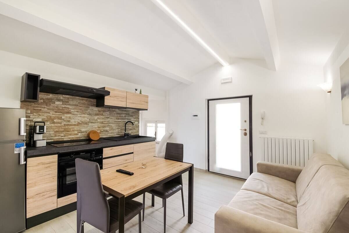A modern kitchen with a wooden countertop, black cabinets, and a built-in stove. There is a dining table with chairs and a beige sofa in the living area.