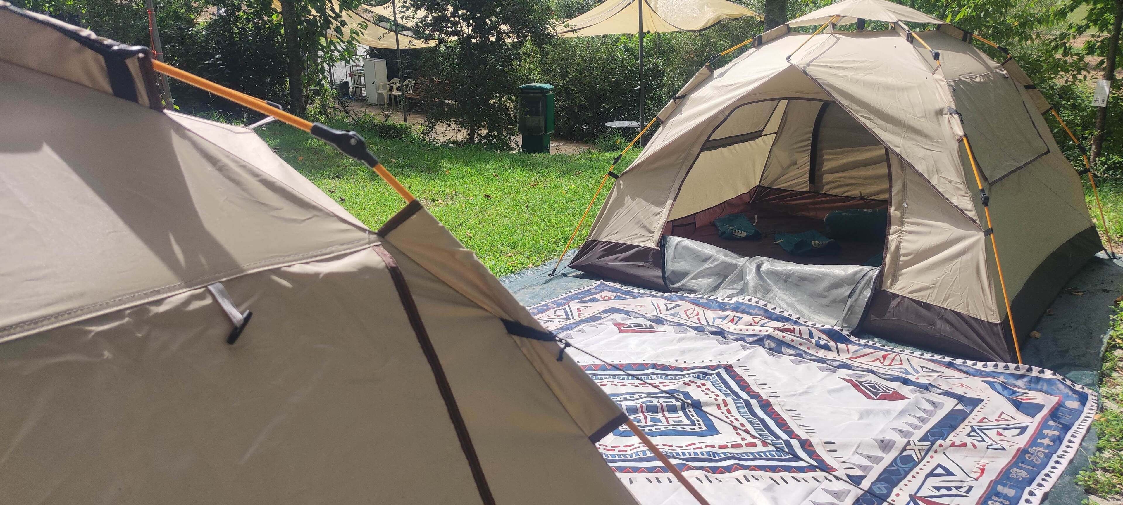 Deux tentes sont installées dans une zone herbeuse, et une couverture à motifs est posée sur le sol.