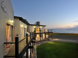 Un bâtiment moderne situé sur la côte, doté d’une terrasse en bois et d’un espace extérieur aménagé, offrant une vue sur une étendue d’eau.