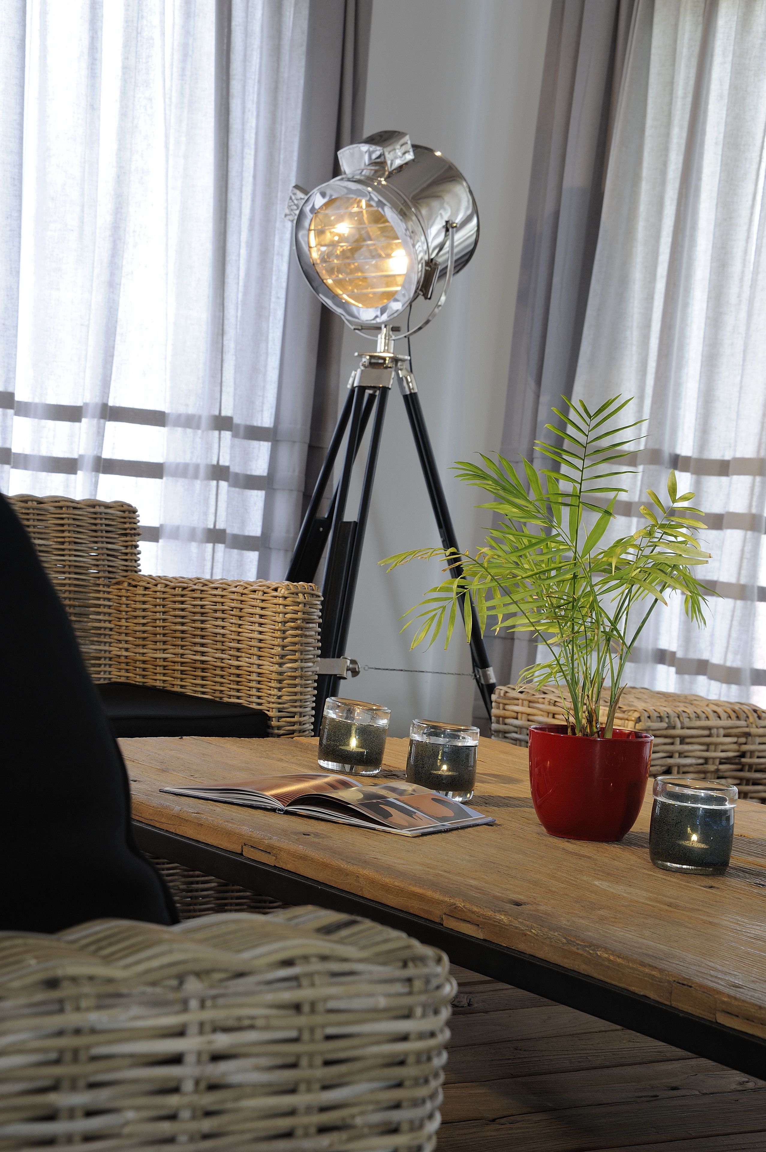 A room with a tripod floor lamp, wicker furniture, a potted plant, and a table with a magazine and glasses.