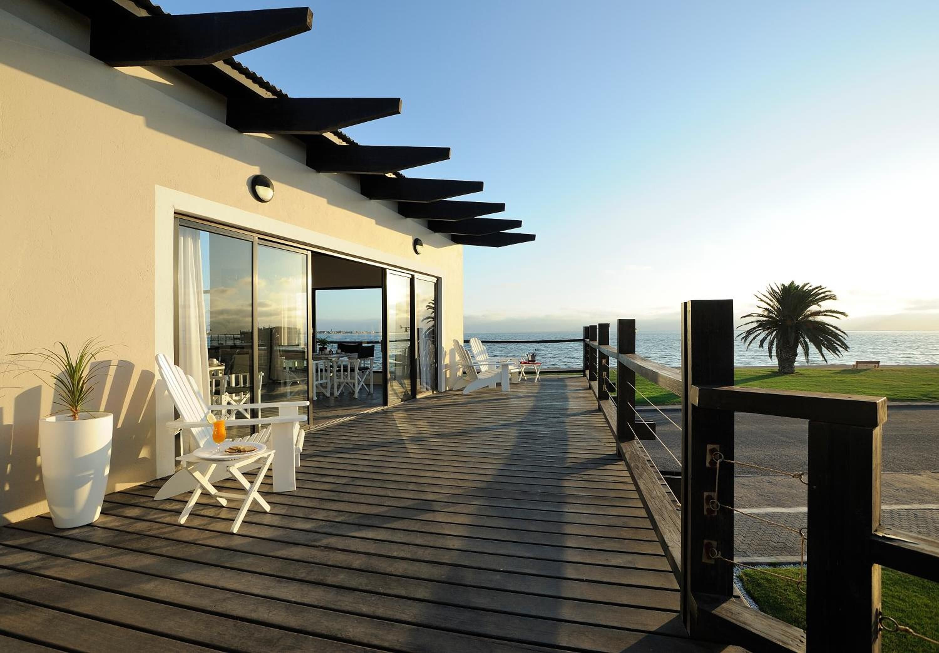 A wooden deck with white chairs and a potted plant, overlooking a body of water.